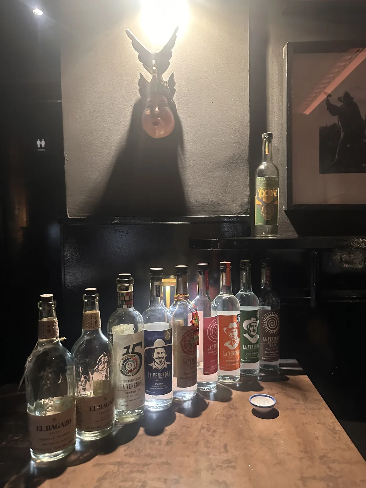 A moody lineup of artisanal mezcal, tequila and raicilla bottles on a dark bar beneath a lit mask.