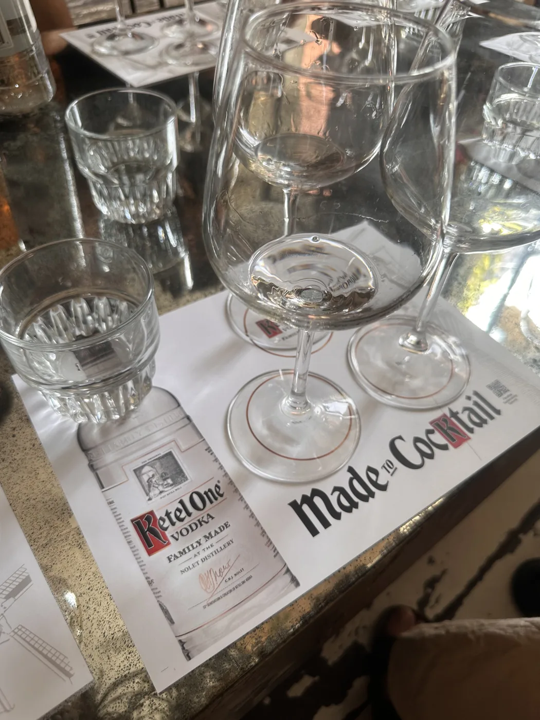 A Ketel One 'Made to Cocktail' tasting sheet with wine glasses and tumblers on the bar.