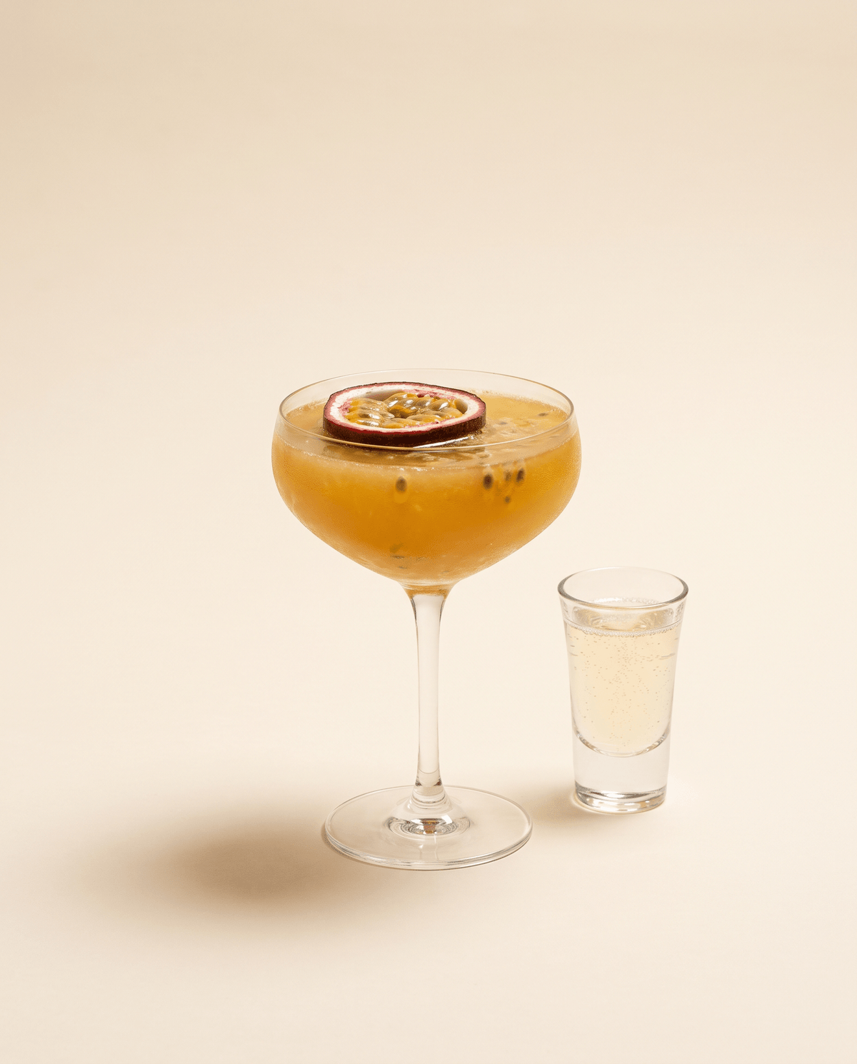 Pornstar Martini — a coupe with a golden passion-fruit cocktail, half a passion fruit floating and a cava shot on the side, on a cream background