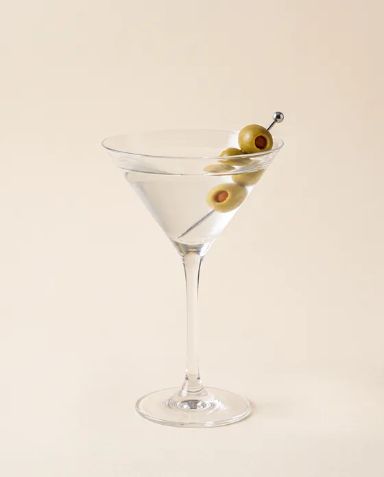 Dirty Martini — a martini glass with a faintly cloudy liquid and olives on a pick, on a cream background