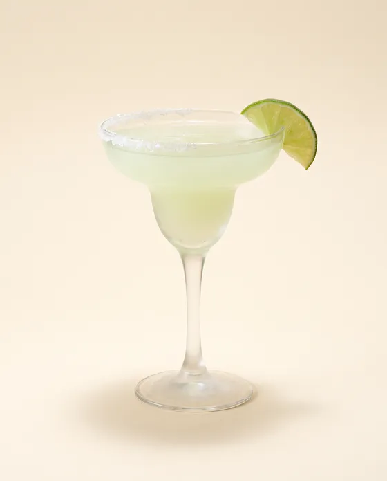 Margarita — a coupe with a pale green-yellow cocktail, a half-moon salt rim and a lime wheel, on a cream background