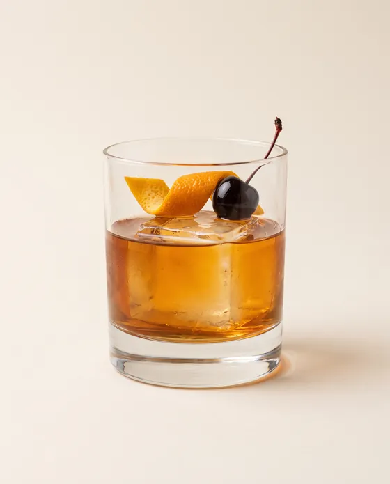 Old Fashioned — a rocks glass with amber whiskey over a large ice cube and an orange peel, on a cream background